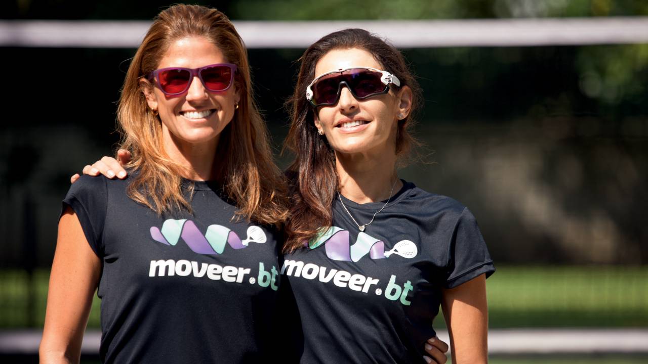 Duas mulheres posam em área ensolarada, abraçadas e sorrindo, com óculos de ciclismo