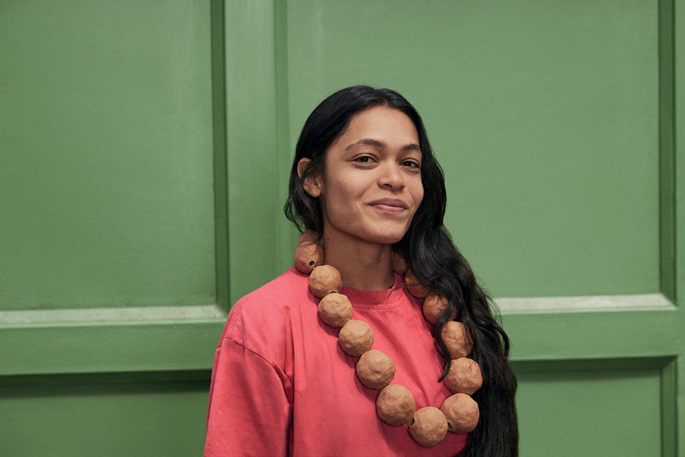Sallisa Rosa portrait _ Cortesia o artista e Audemars Piguet Contemporay.jpg Mulher indígena de cabelos pretos longos sorri olhando para a foto, usa colar grande de esferas marrons e usa peça rosa