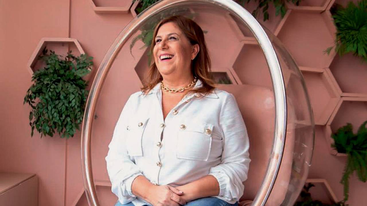 Mulher branca de cabelos lisos posa sentada sorrindo e olhando para a esquerda em cadeira oval transparente. Ao fundo, parede cor de salmão com plantas