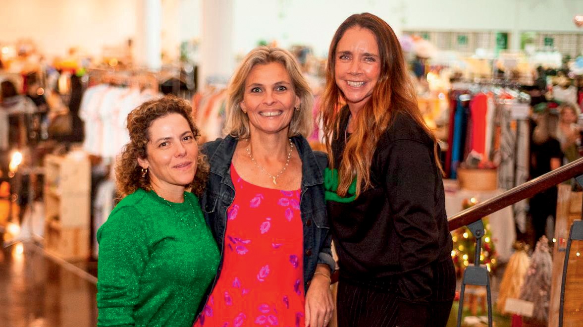Três mulheres posam lado a lado em loja. A partir da esquerda, vestem peça verde, rosa e preta. As três sorriem.