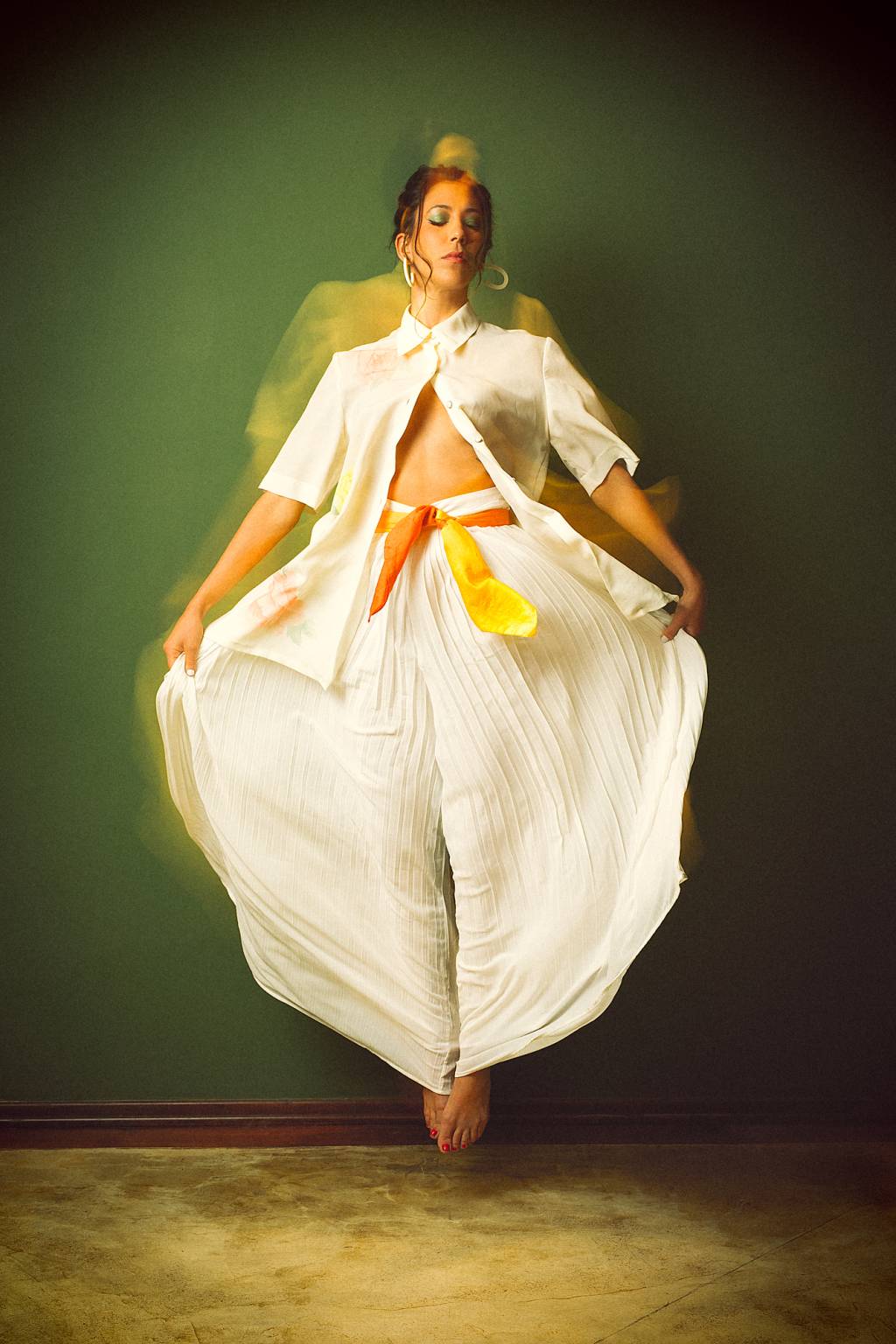 Liza Schechtmann-Divulgação (3) Mulher branca de cabelos escuros curtos está puxando o tecido de uma longa calça branca e veste camisa branca com brincos claros. O fundo é verde.