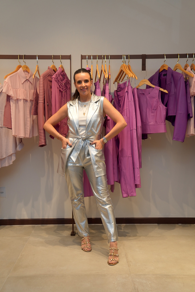 Mariana Orlando-Crédito Fábio Ponce-Divulgação (5).jpg Mulher branca vestida com peças prateadas posa com mãos na cintura em frente a arara com peças em tons de rosa