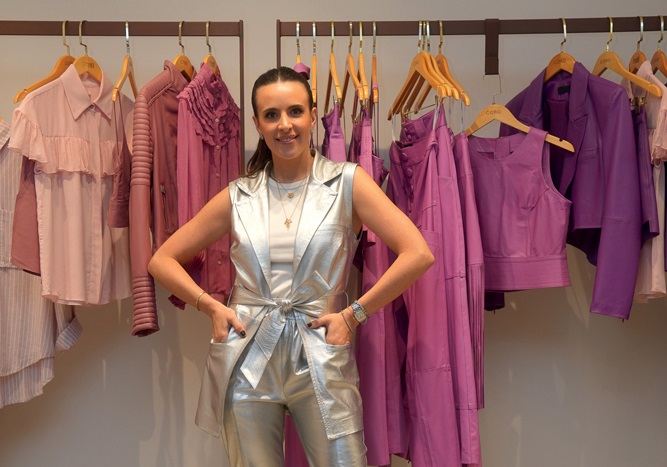 Mulher branca vestida com peças prateadas posa com mãos na cintura em frente a arara com peças em tons de rosa