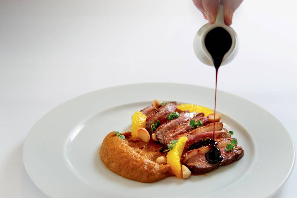 Bistrot du Quartier Prato de louça branca com purê alaranjado ao lado de fatias ao ponto de carne sendo cobertas por molho saindo de jarrinha