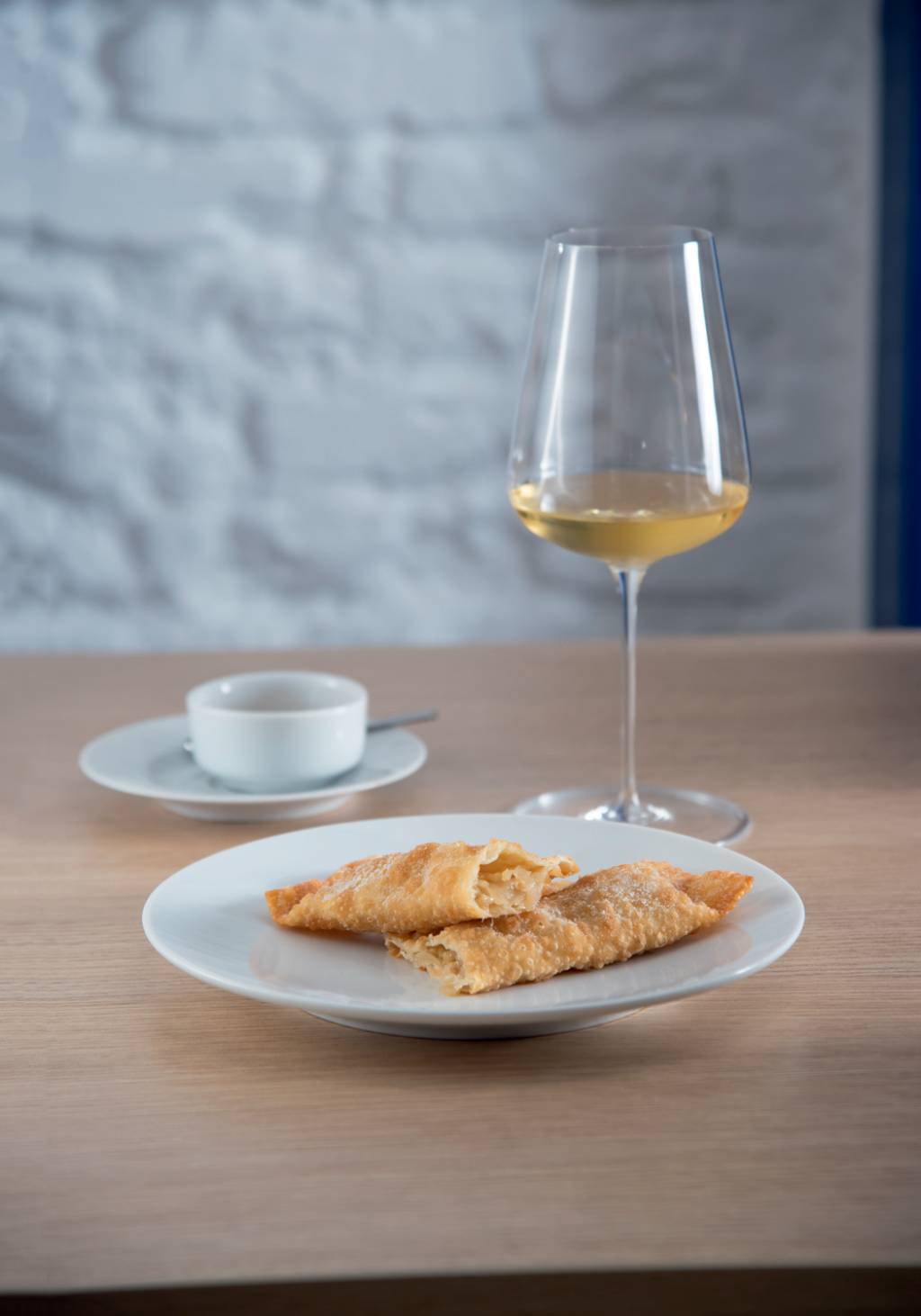 Cellar Cave Prato de louça branca com dois pastéis retangulares sobre mesa de mandeira em frente a taça com vinho branco