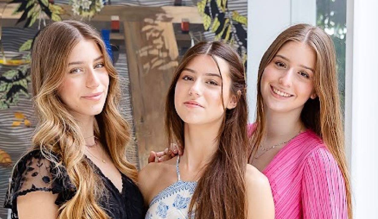 Três meninas brancas de cabelos longos lisos sorriem para a câmera, a partir da esquerda vestindo peças nas cores preta, branca e rosa