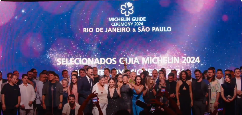 Print de vídeo com dezenas de pessoas em cima de palco em frente a telão escrito "MIchelin Guide Rio de Janeiro & São Pualo".