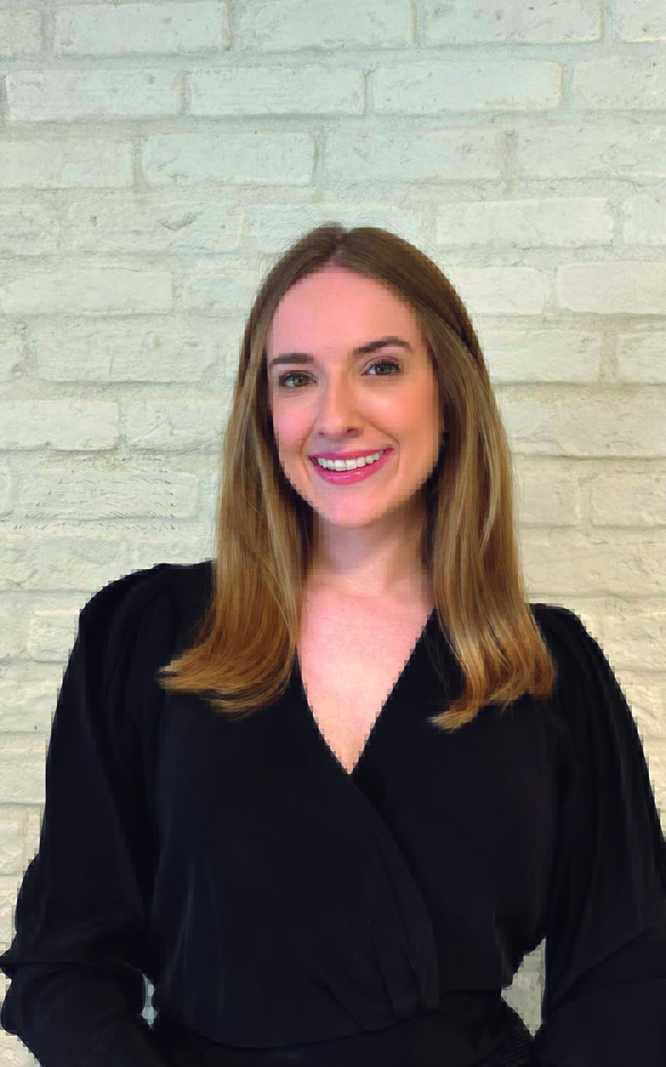 Bruna Soares, head da Veuve Clicquot no Brasil Mulher loira sorri vestindo peça preta e posando em fundo branco de tijolos