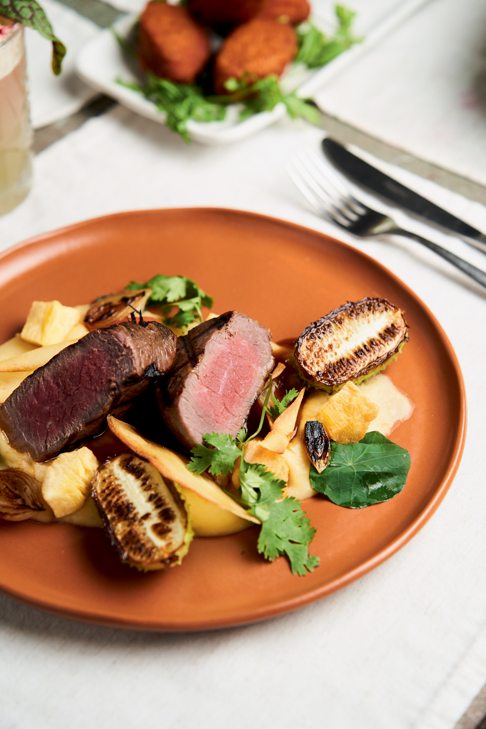 Dia dos Namorados – Preto Cozinha Prato de cerâmica laranja com creme de mandioca coberto por fatias de carne rosada, pedaços de maxixe tostada e folhas de coentro.
