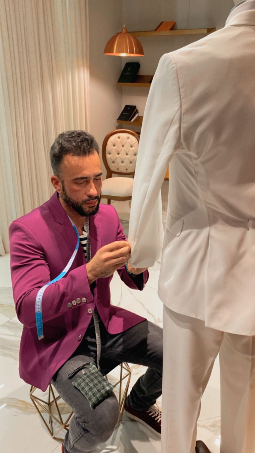 Charles Hermann nova coleção masculina (1) Homem de terno roxo posa agachado mexendo em terno branco, com fita métrica no antebraço