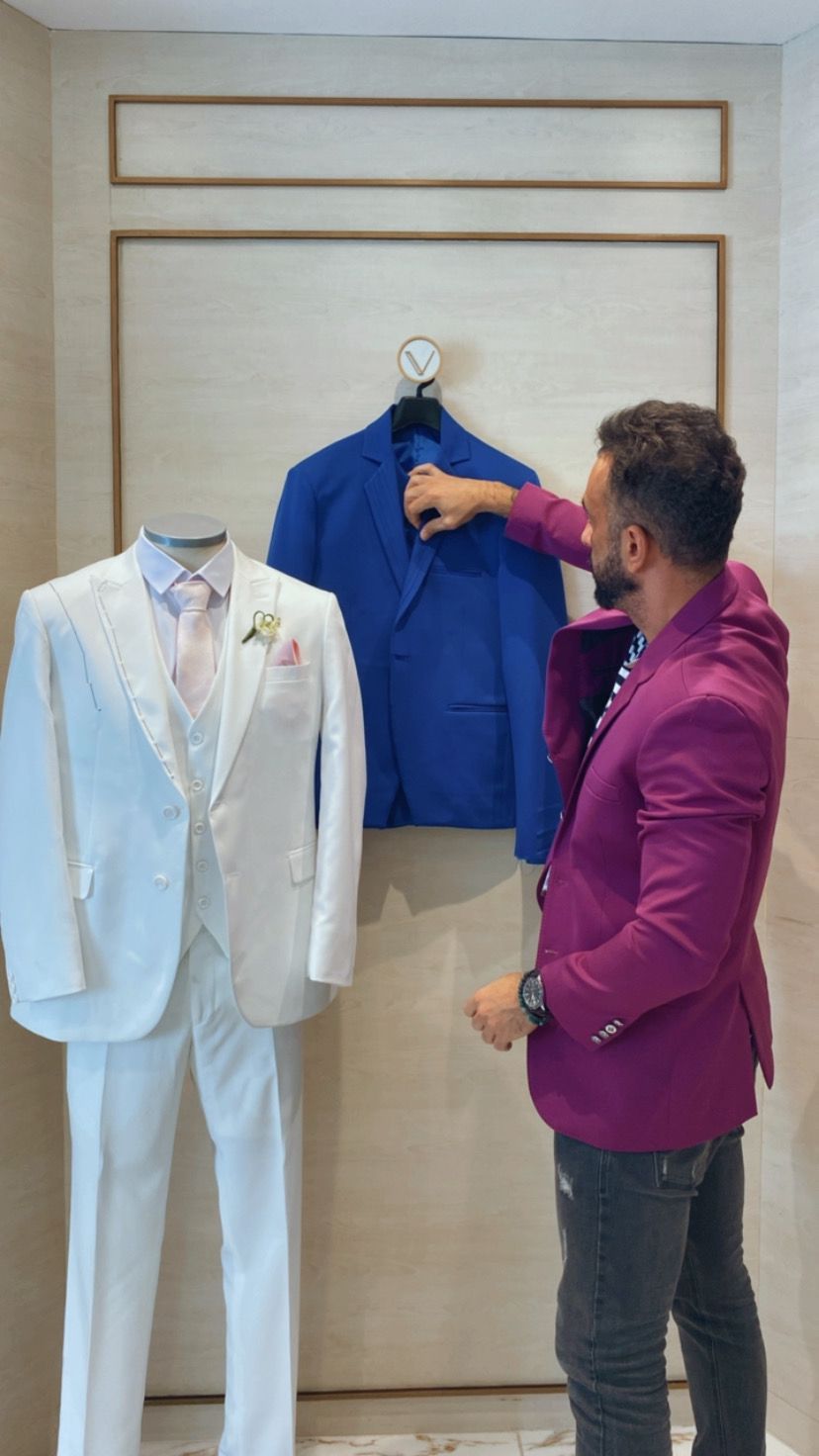 Charles Hermann nova coleção masculina (2) Homem de terno roxo mexe em paletó azul pendurado na parede. À esquerda tem um smoking completo branco em exposição