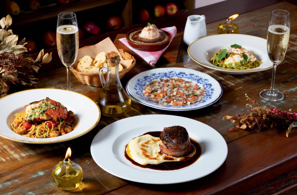 Dia dos Namorados – Osteria Nonna Rosa Mesa de madeira com diversos pratos e duas taças de espumante. À frente, prato raso de louça branca com medalhão ao lado de aligot, à esquerda macarrão em prato fundo coberto por lagosta, ao centro prato branco com detalhes em azul e vinagrete, à direita, macarrão verde com creme branco.