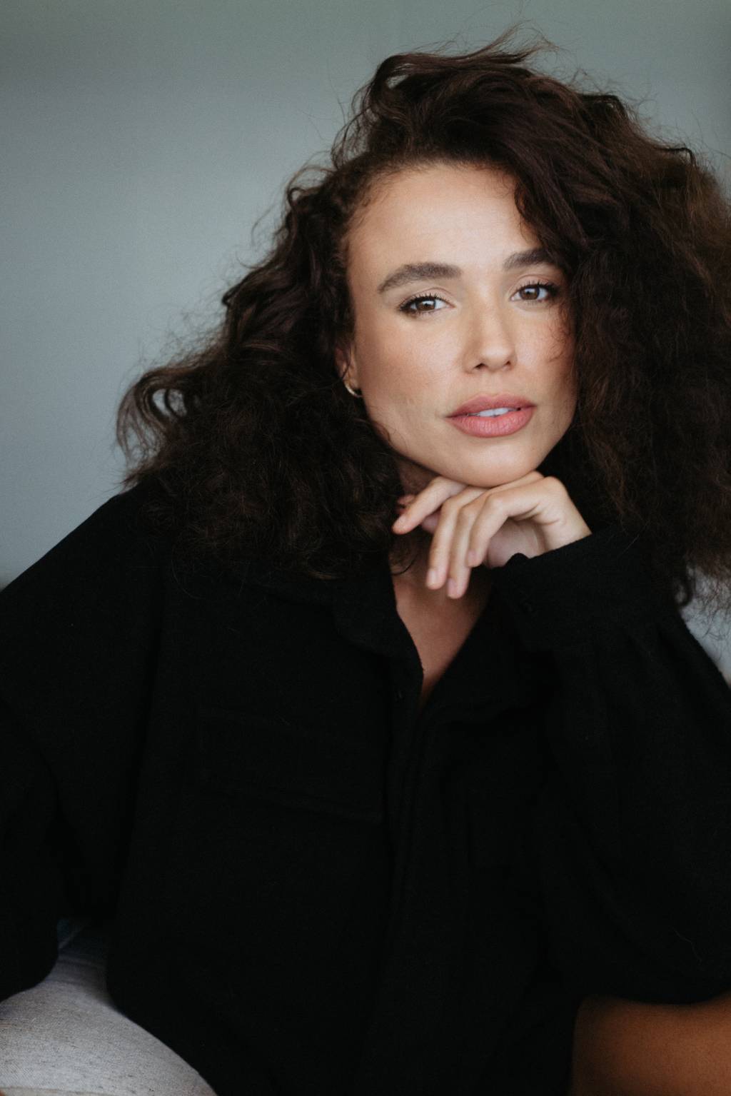 Juliane Araújo Moça branca de cabelos escuros volumosos posa com mão no rosto
