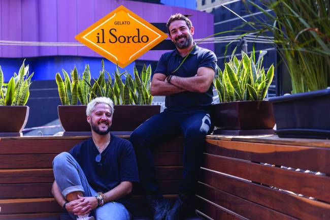 André Vasco e Felipe Paladino _ il Sordo Os dois homens sentados num banco de madeira na frente da placa da sorveteria