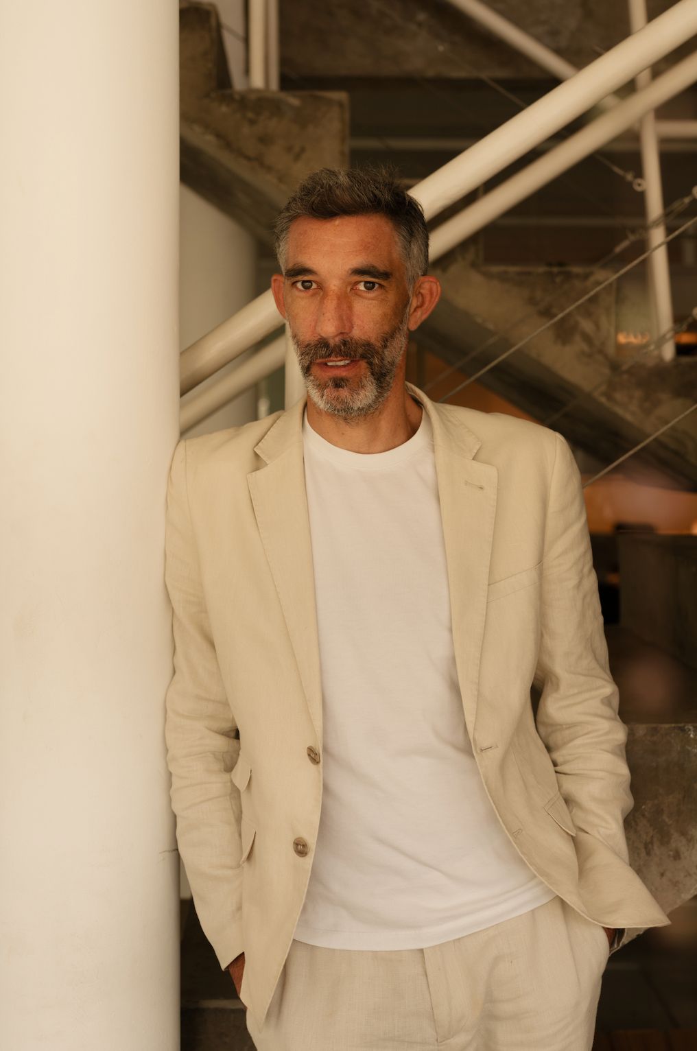 Greg Bousquet Homem alto e branco de barba posa encostado em pilastra branca vestindo roupas brancas e paletó bege claro
