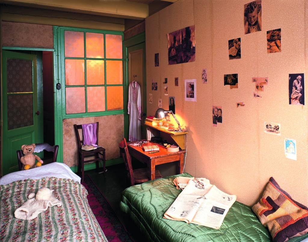 Kamer Anne en Pfeffer ingericht_Coleção de fotos da Anne Frank Stichting (Amsterdam) Crédito Acervo Anne Frank House-Reprodução Foto de quarto com cama verde, porta verde com vidros, outra cama à esquerda e fotos na parede branca