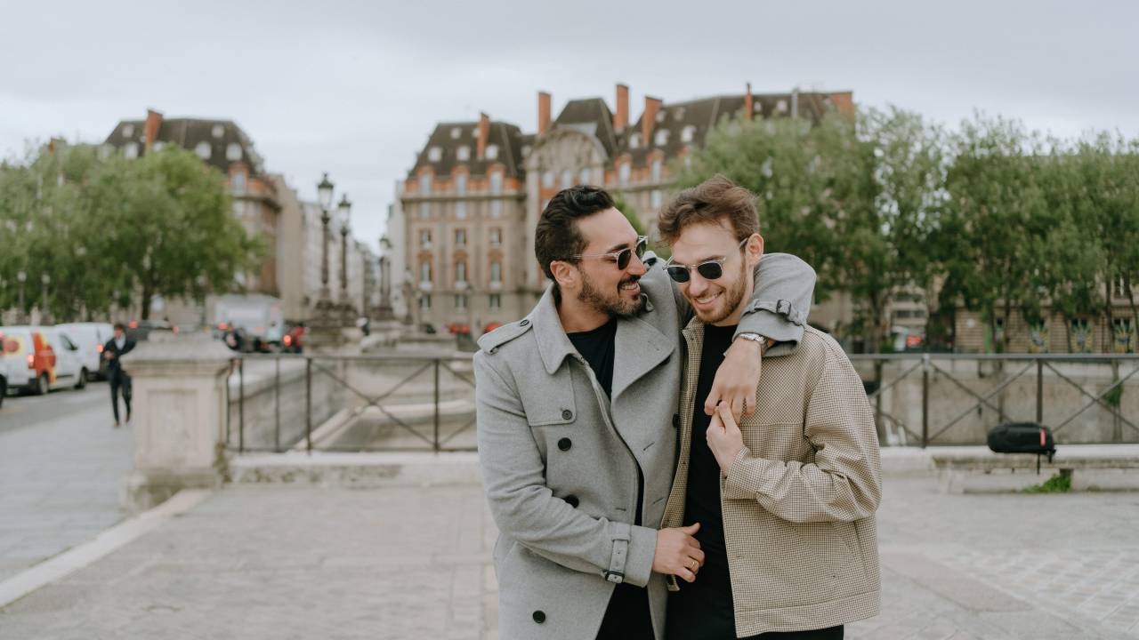 Dois moços brancos sorriem abraçados em rua de Paris, ambos de óculos escuros e roupas de frio