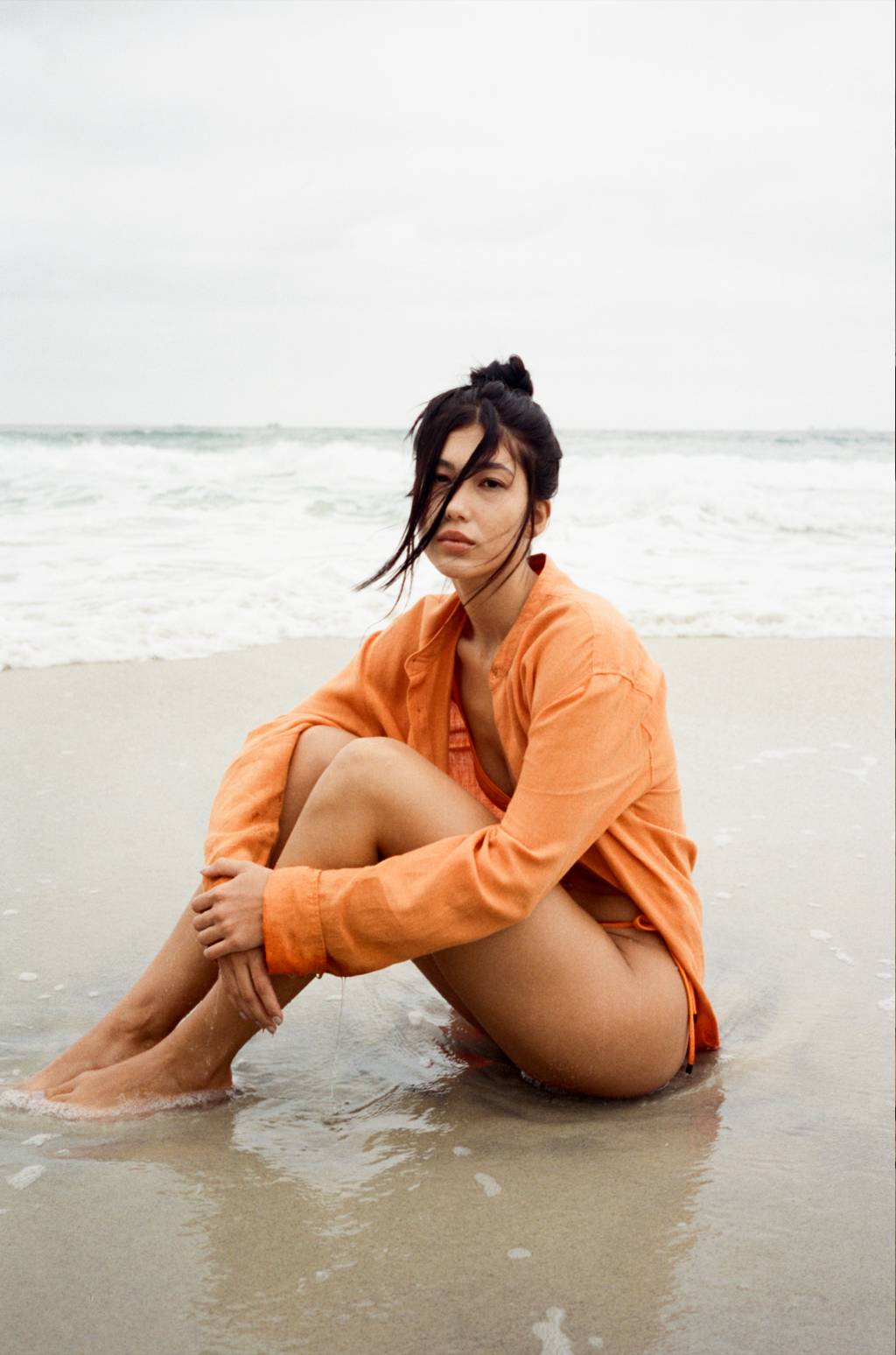 Osklen coleção verão 2025 (1) Moça asiática sentada na areia com roupa laranja