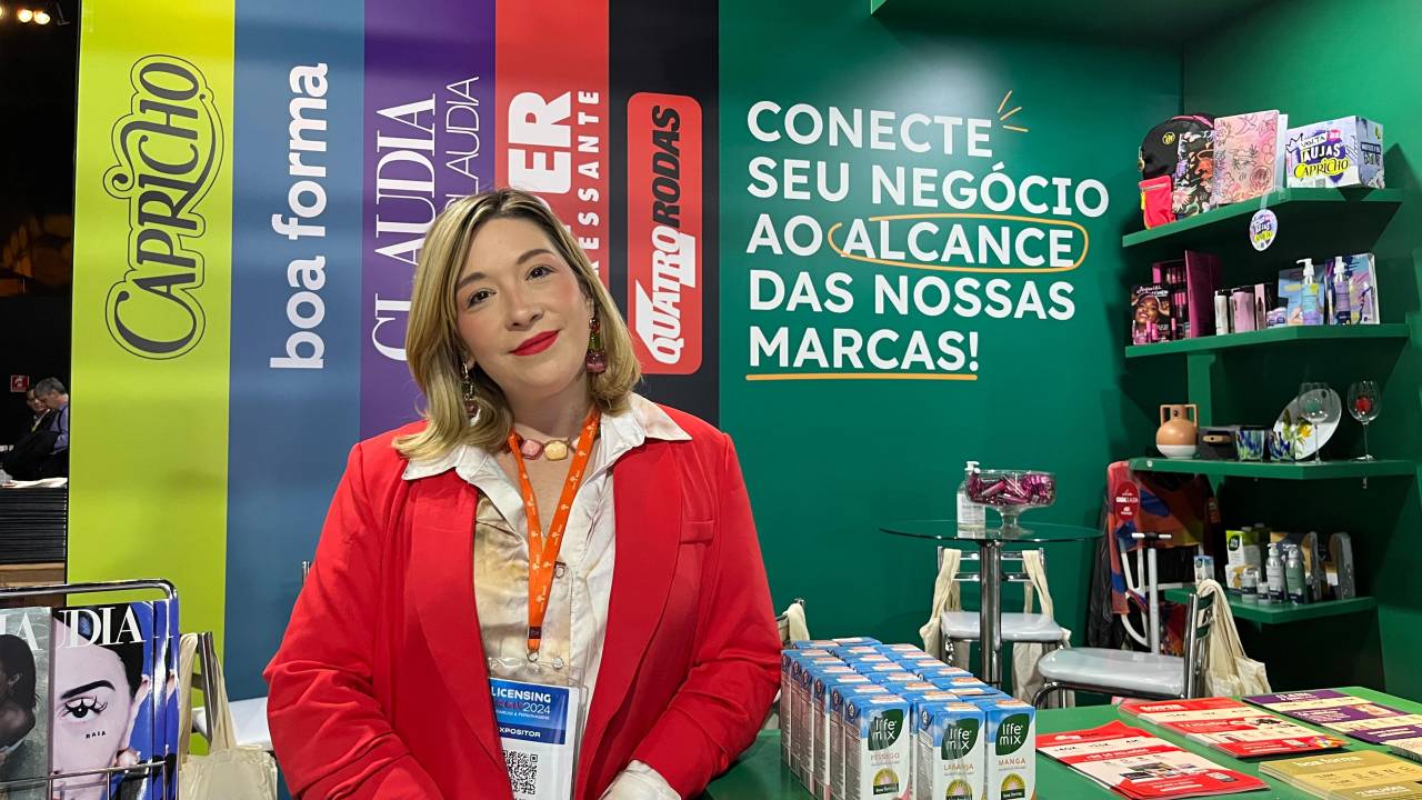 Mulher loira de terno vermelho posa em frente a estande verde com marcas de revista anunciadas