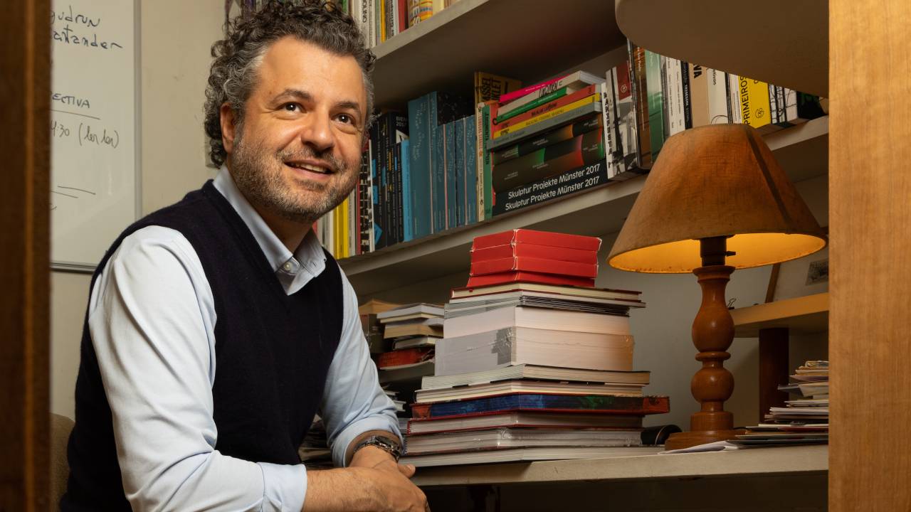Homem branco de cabelo cinza posa sentado ao lado de pilha de livros