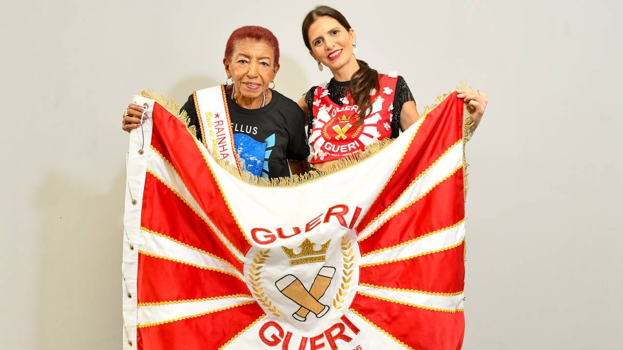 Senhora e moça posam segurando bandeira vermelha e branca de bloco carnavalesco