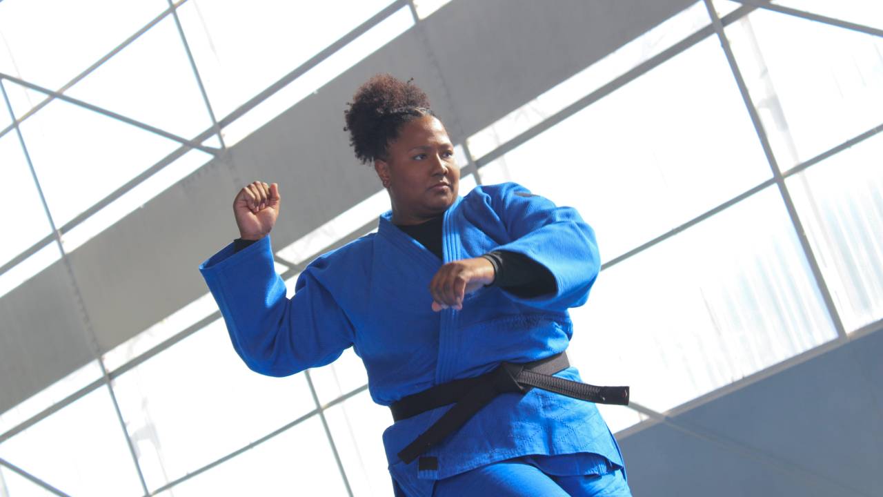 Mulher negra ergue o punho direito em tom de comemoração. Veste quimono azul com faixa preta.