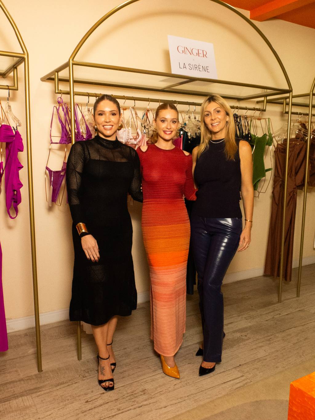 Carol Gazoto, Marina Ruy Barbosa e Michelle Coleto Crédito Ana Candreva Photo-Divulgação. (2) Três mulheres posam em frente a arara de roupas em loja. A do meio, ruiva, usa vestido laranja em tons degradê. As loiras nas duas pontas estão de preto.