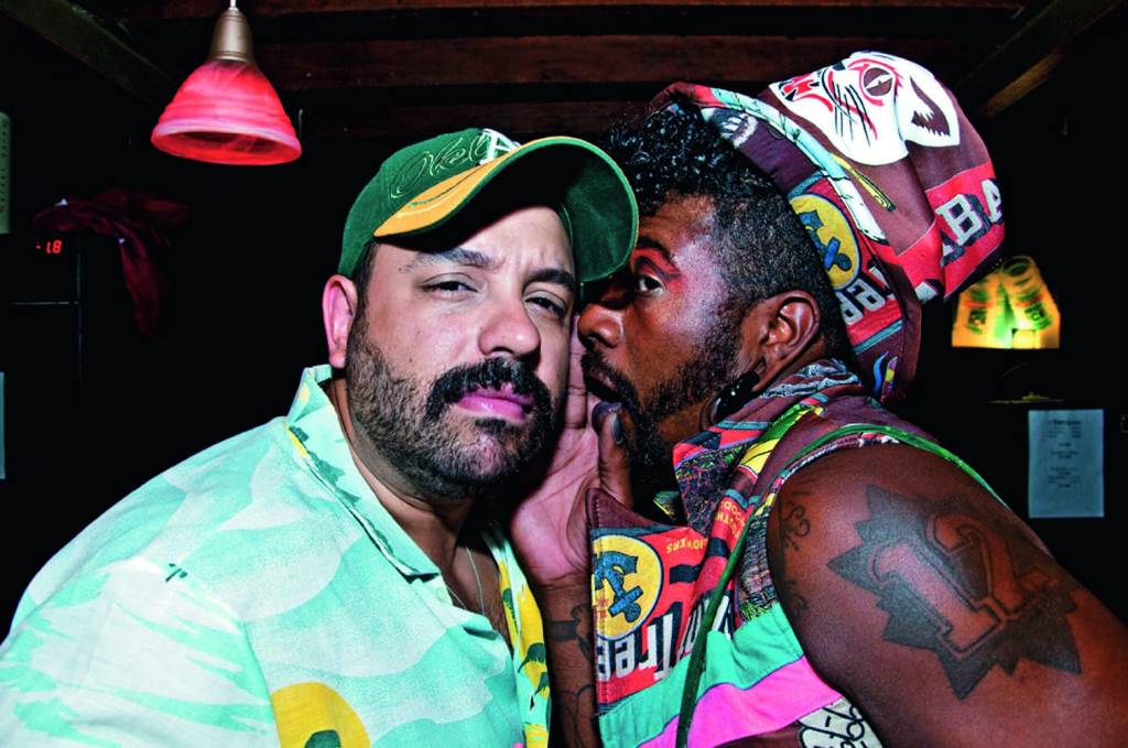 DJ Bispo e Jota Jota Davis Crédito Paulo Batalha-Reprodução Homem branco de barba e homem negro à direita perto de seu ouvido, o da esquerda usa boné e o da direita adereços coloridos no pescoço