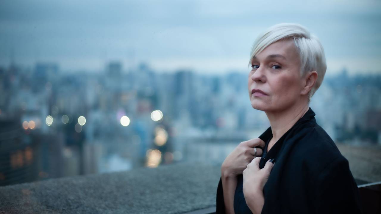 Moça loira de cabelo curto posa com blazer preto e cidade do alto aparece ao fundo
