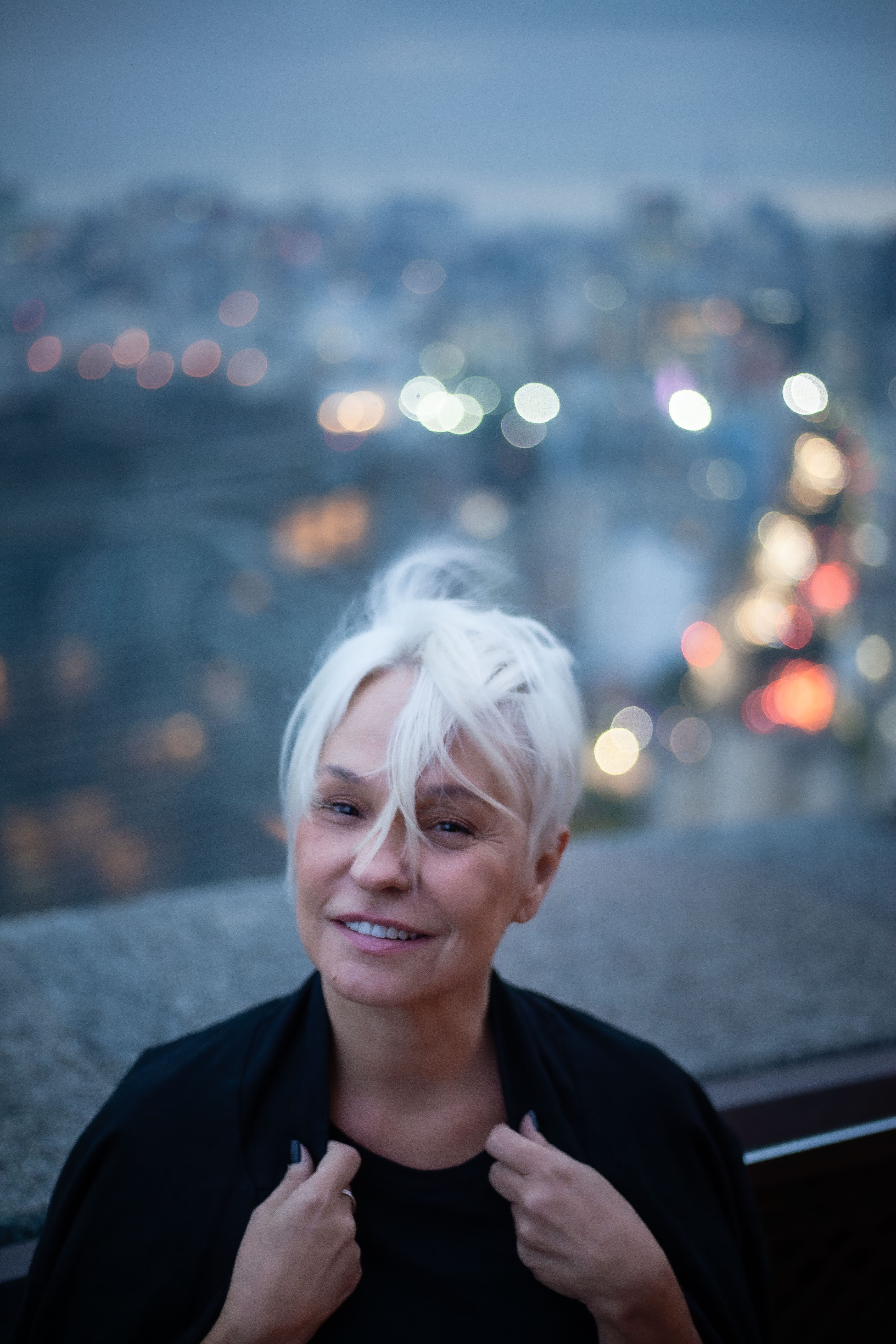 Mo&ccedil;a loira de cabelo curto posa com blazer preto e cidade do alto aparece ao fundo