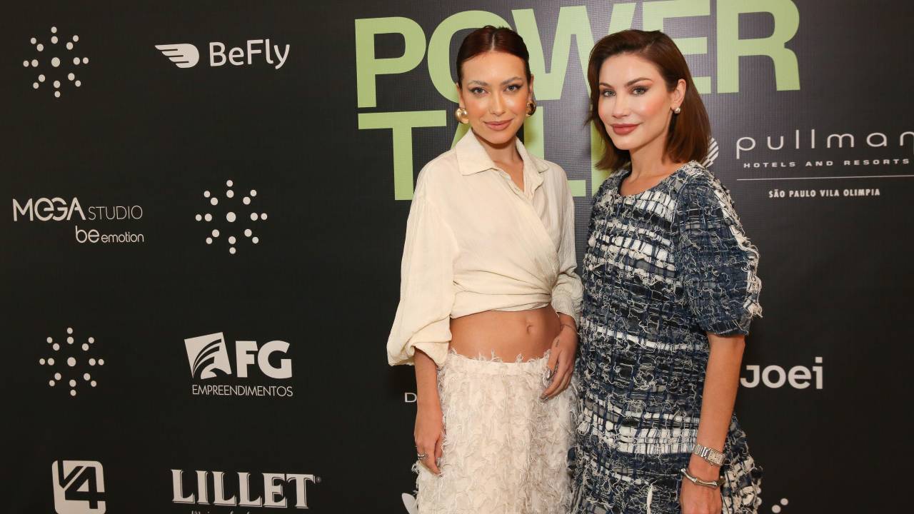 Dupla de mulheres posam em evento com fundo preto e estampas de marcas
