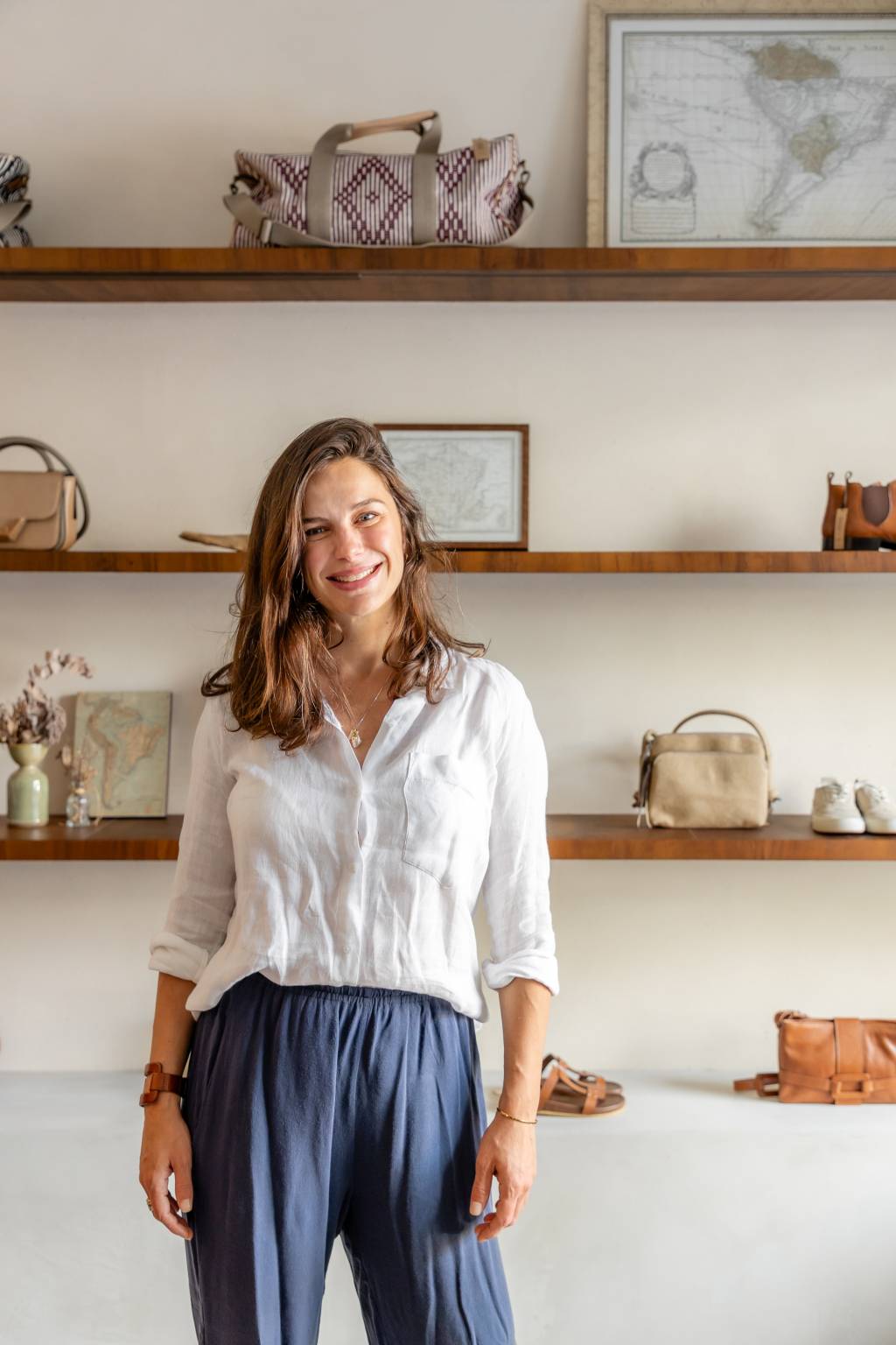 Manuela Rodrigues, Cabana Crafts Crédito Ricardo Faiani-Divulgação (1) Moça branca de camisa branca e calça azul posa em frente a prateleiras de utensílios de casa