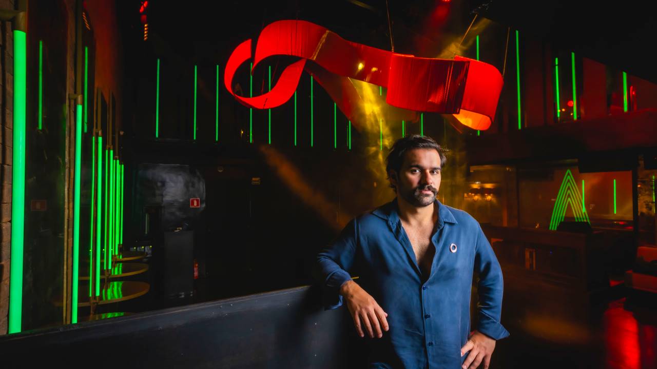 Homem de barba e camisa azul apoiado em balcão preto em frente a balada com ornamento vermelho no teto