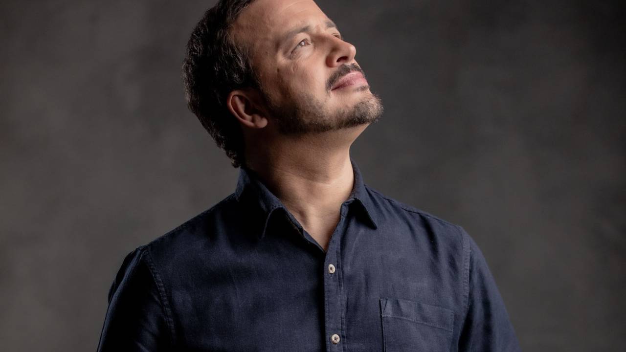 Homem branco posa de camisa azul curta olhando para cima com olhar pensativo