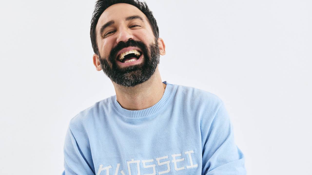 Homem de barba preta veste moletom azul bebê, calça preta, e posa sentado rindo