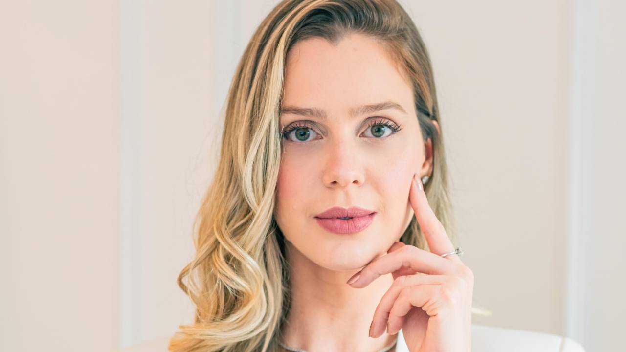 Doutora de jaleco e camiseta azul, mão no queixo, cabelo loiro longo e liso