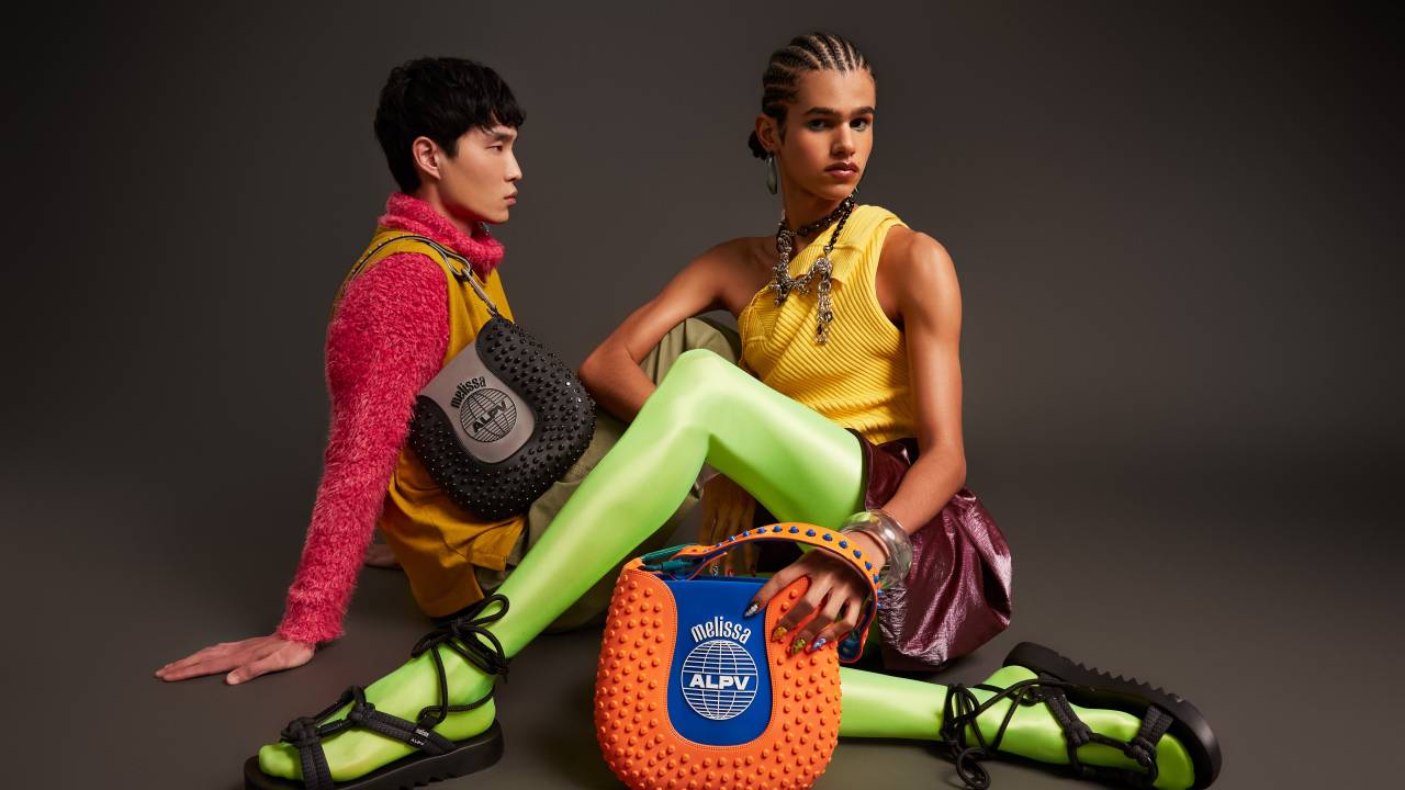 Foto tem dois modelos com roupas coloridas e uma grande bolsa no meio