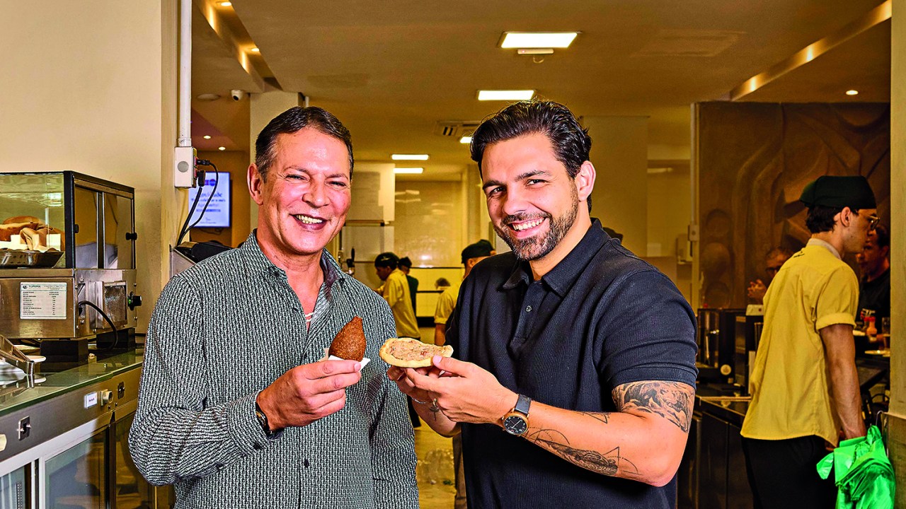 Dois homens sorrindo para foto e segurando salgados