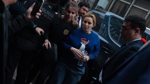 Mulher loira de suéter azul com urso estampado, segurando uma garrafa d'água, sendo escoltada por uma policial civil e outros homens em meio a uma multidão
