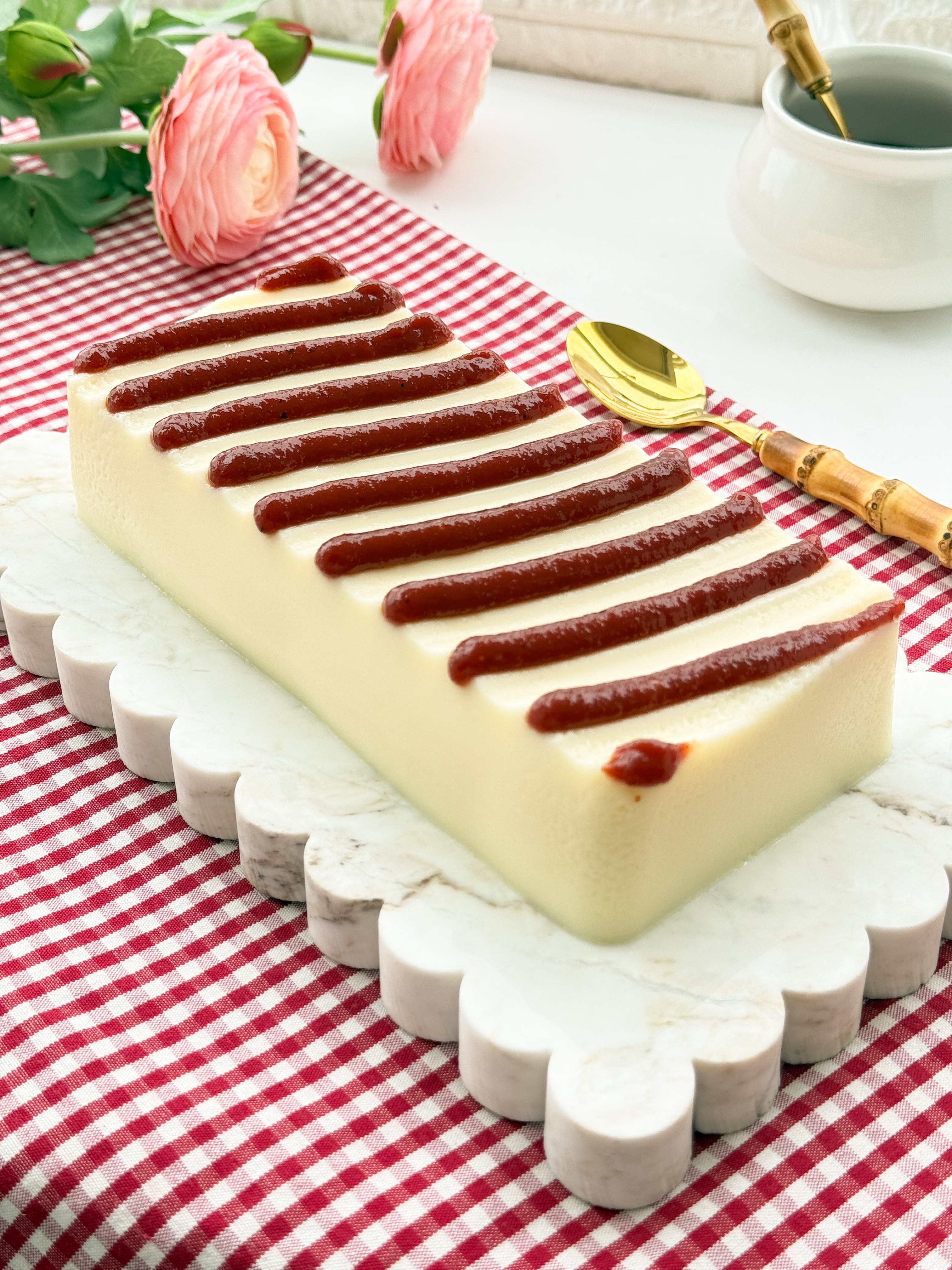 Bolo de queijo cremoso retangular com tiras de goiabada por cima, servido em uma t&aacute;bua branca decorada, sobre uma toalha xadrez vermelha e branca. Ao fundo, flores cor-de-rosa e uma colher dourada com cabo de bambu