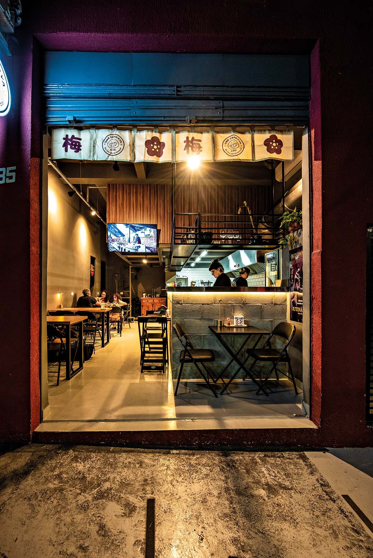 Fachada de um restaurante japon&ecirc;s &agrave; noite, com cortinas noren brancas e caracteres orientais. Dentro, clientes sentados em mesas de madeira, balc&atilde;o de tijolos com cozinheiros e uma televis&atilde;o exibindo imagens. A ilumina&ccedil;&atilde;o quente cria um ambiente acolhedor