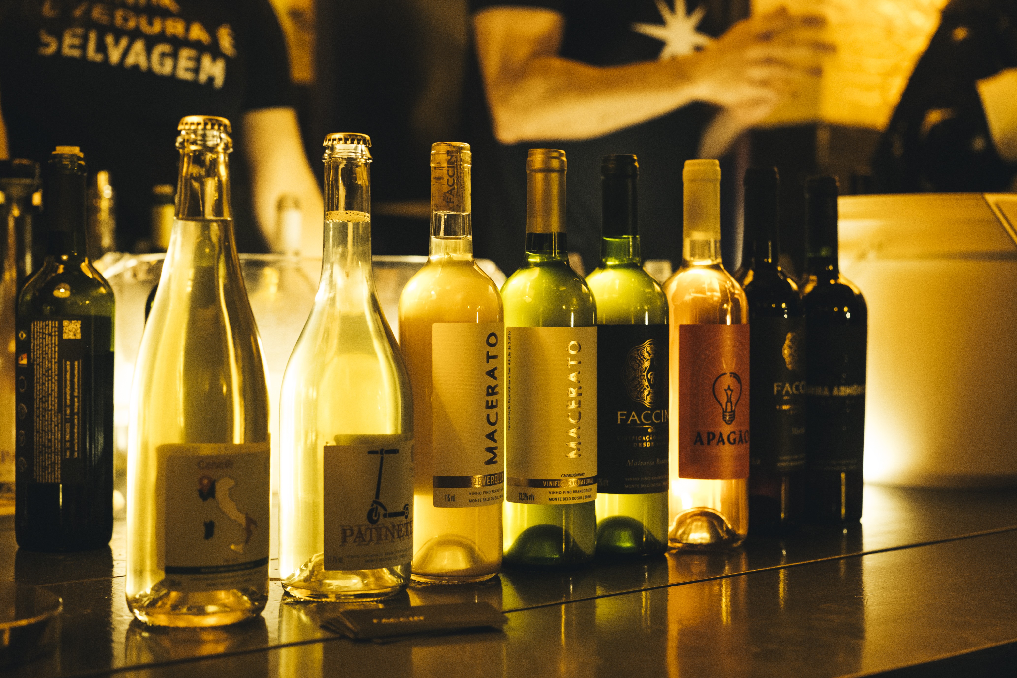 Garrafas de vinho e espumante em tons de verde, &acirc;mbar e transparente, alinhadas sobre um balc&atilde;o escuro e brilhante, com ilumina&ccedil;&atilde;o quente ao fundo, onde uma pessoa de camiseta preta com a frase SELVA E SELVAGEM &eacute; vis&iacute;vel