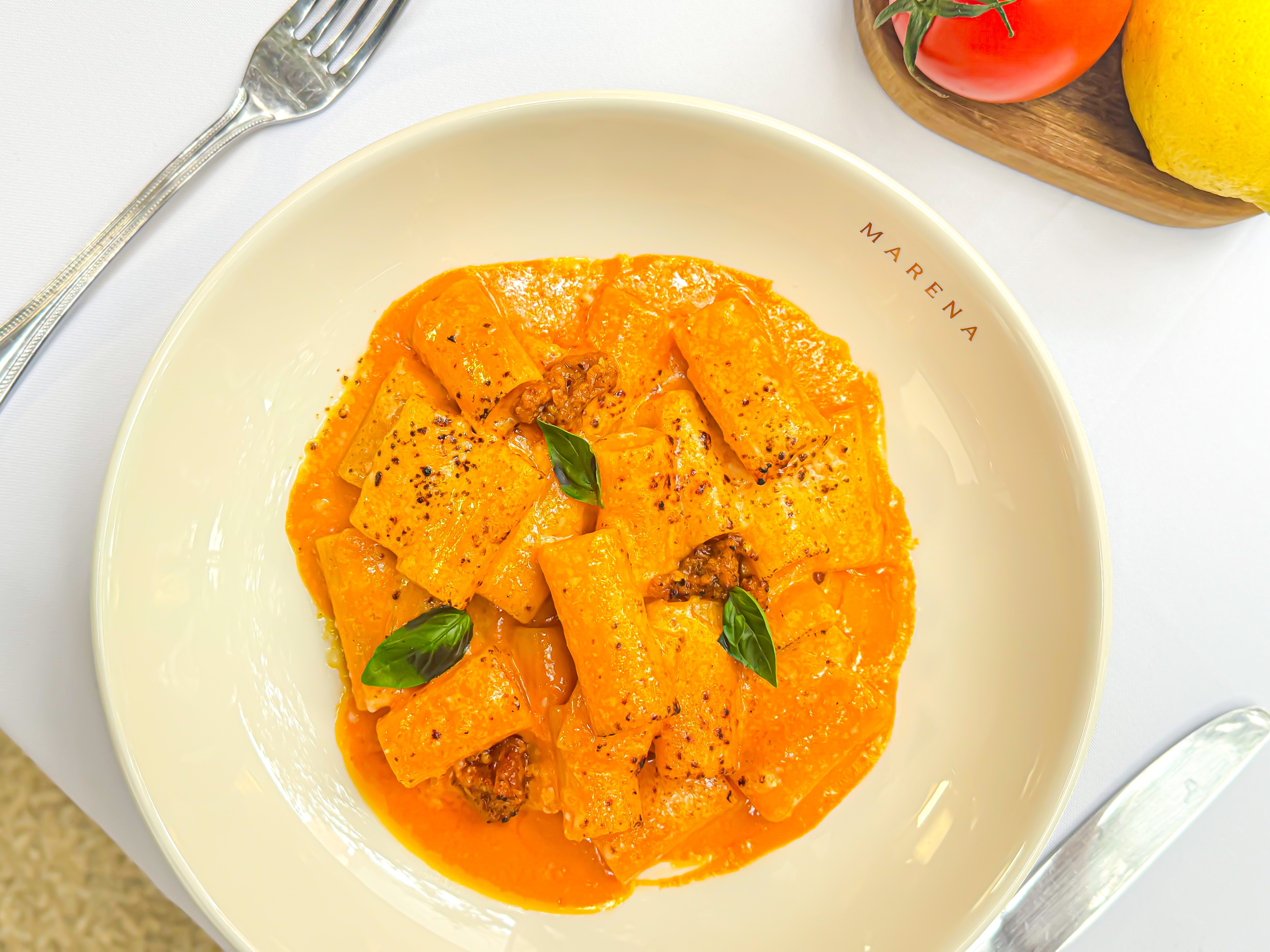 Prato de massa rigatoni com molho cremoso laranja, salpicado com pimenta preta e folhas de manjeric&atilde;o fresco, em um fundo branco com talheres e ingredientes
