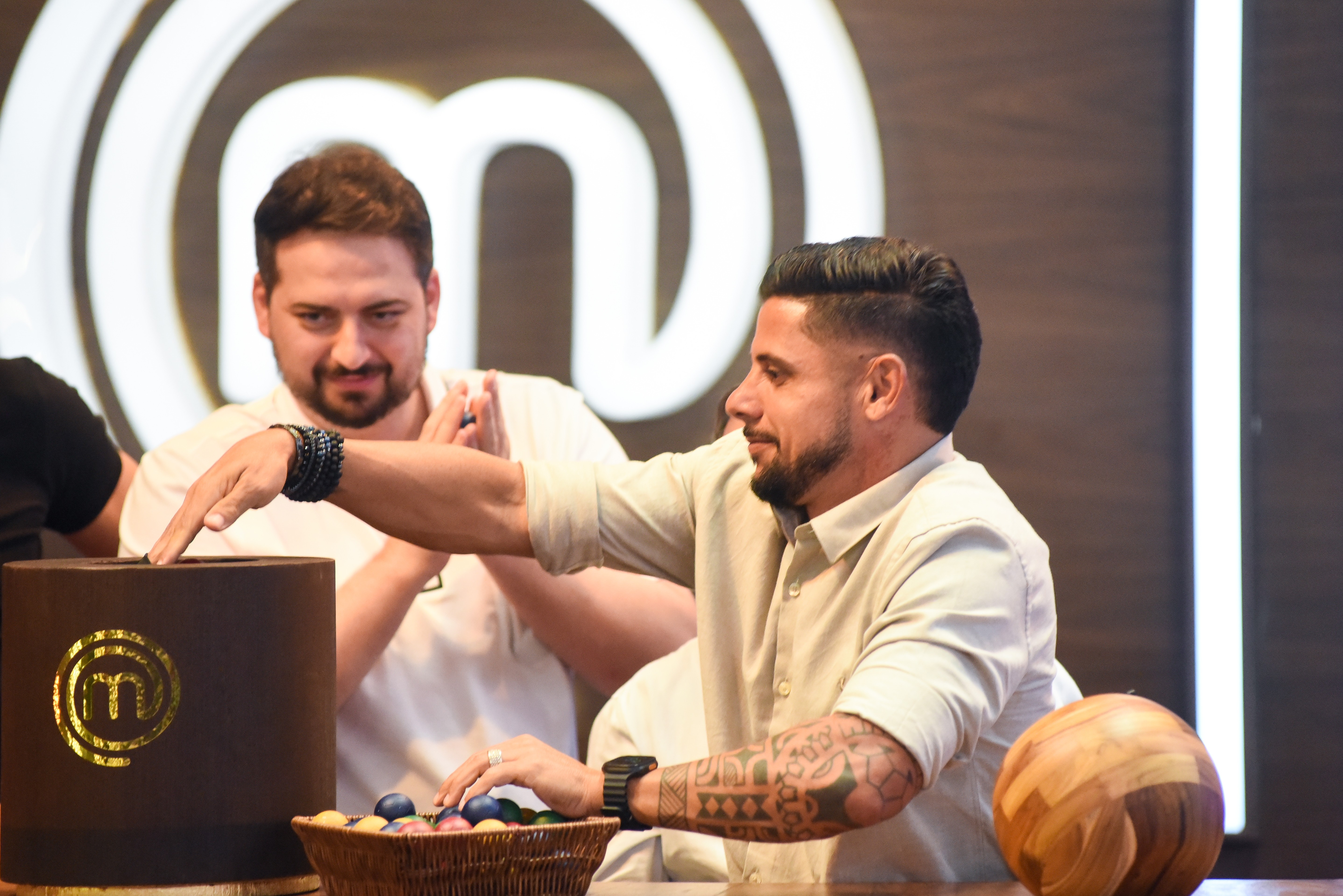 Dois homens em um evento. Um homem barbudo de camisa bege e bra&ccedil;os tatuados pega algo de uma cesta com ovos coloridos. Outro homem barbudo de camisa branca sorri ao fundo, com a m&atilde;o sobre uma caixa marrom com o logo 