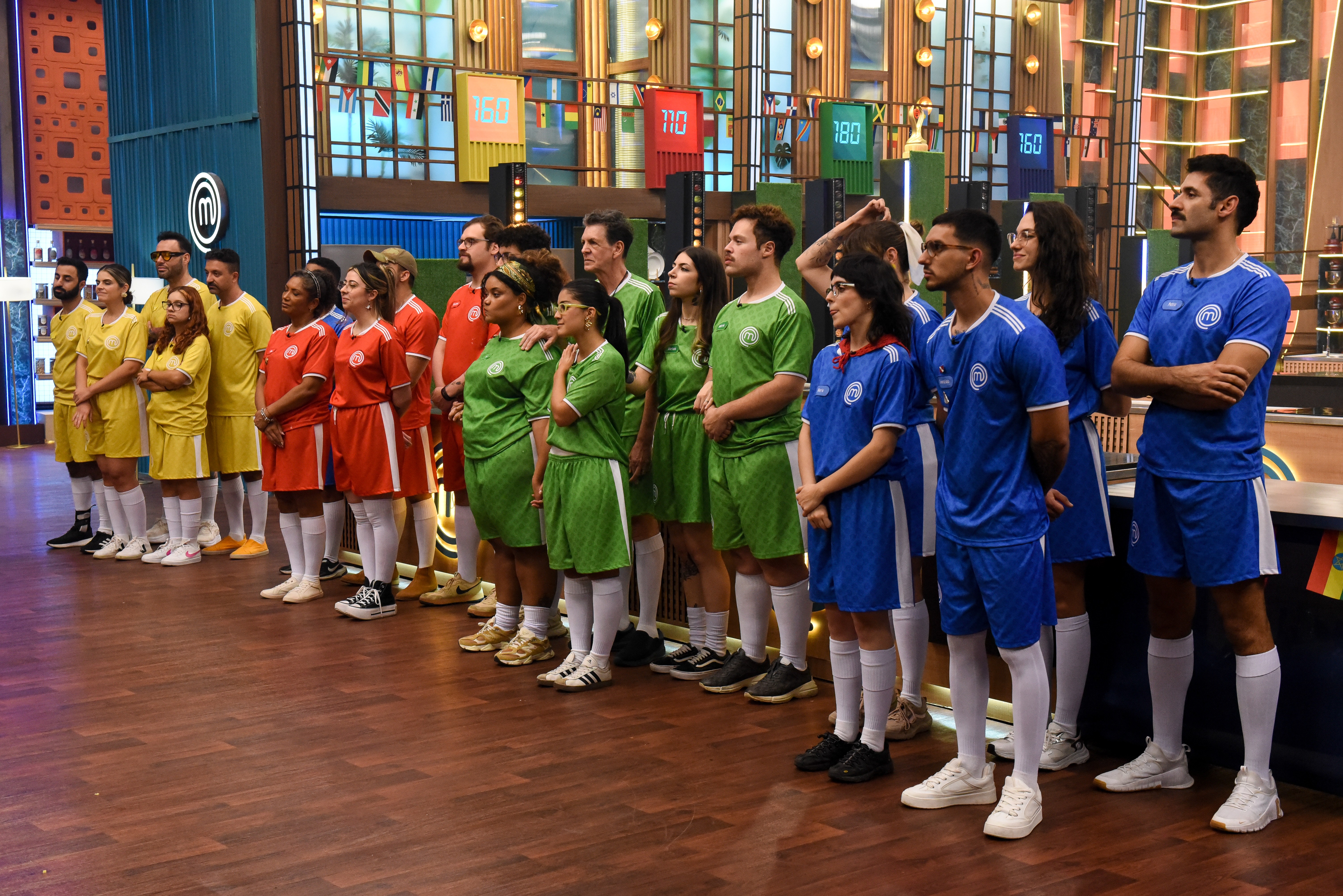 Grupo de 20 pessoas, homens e mulheres, em p&eacute;, alinhadas em um est&uacute;dio de TV. Usam uniformes esportivos de futebol nas cores azul, verde, vermelho e amarelo, com meias brancas e t&ecirc;nis. Ao fundo, um cen&aacute;rio com pain&eacute;is coloridos e luzes.