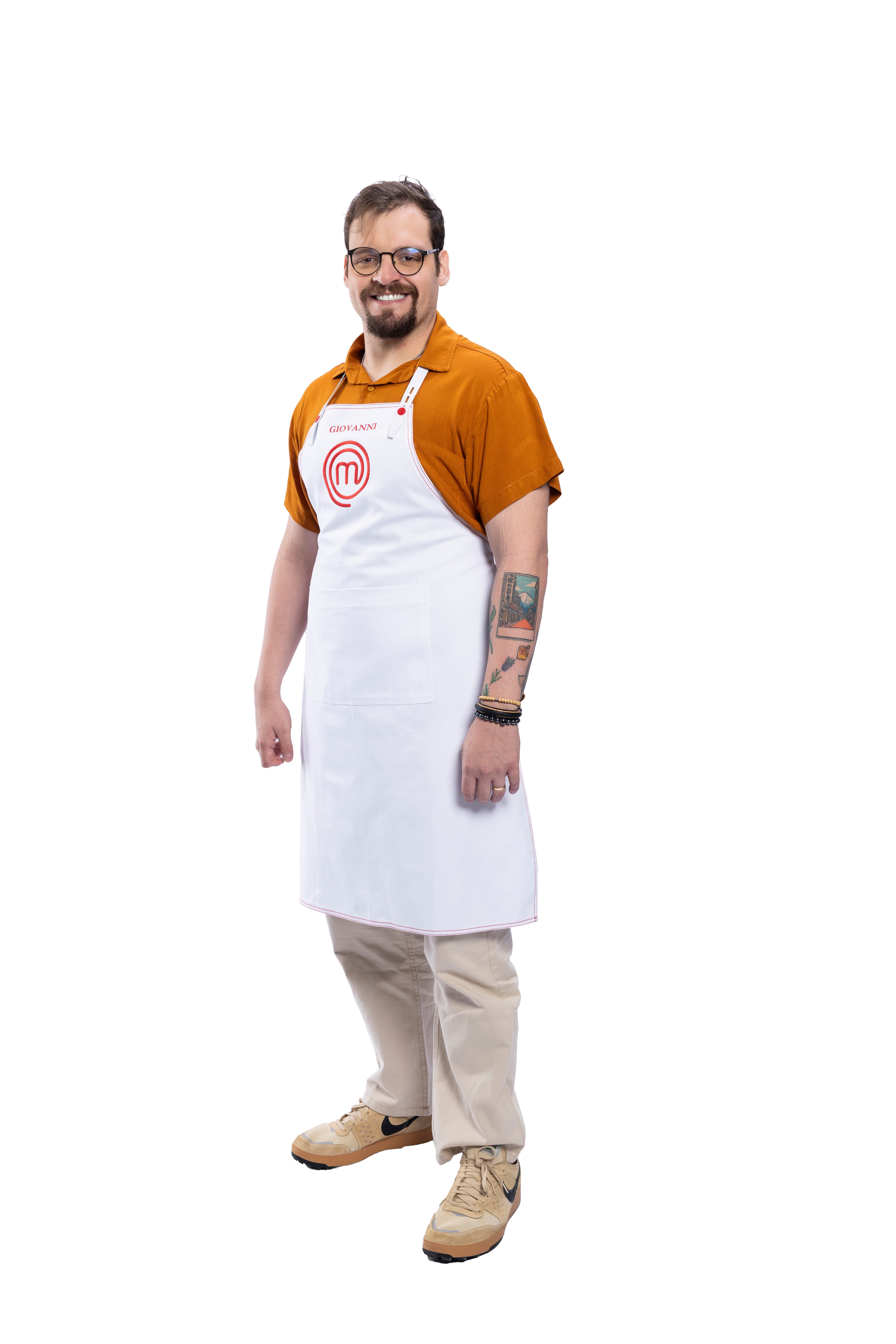 Homem barbudo, de &oacute;culos, sorrindo, vestindo camisa laranja, avental branco do MasterChef com o nome 