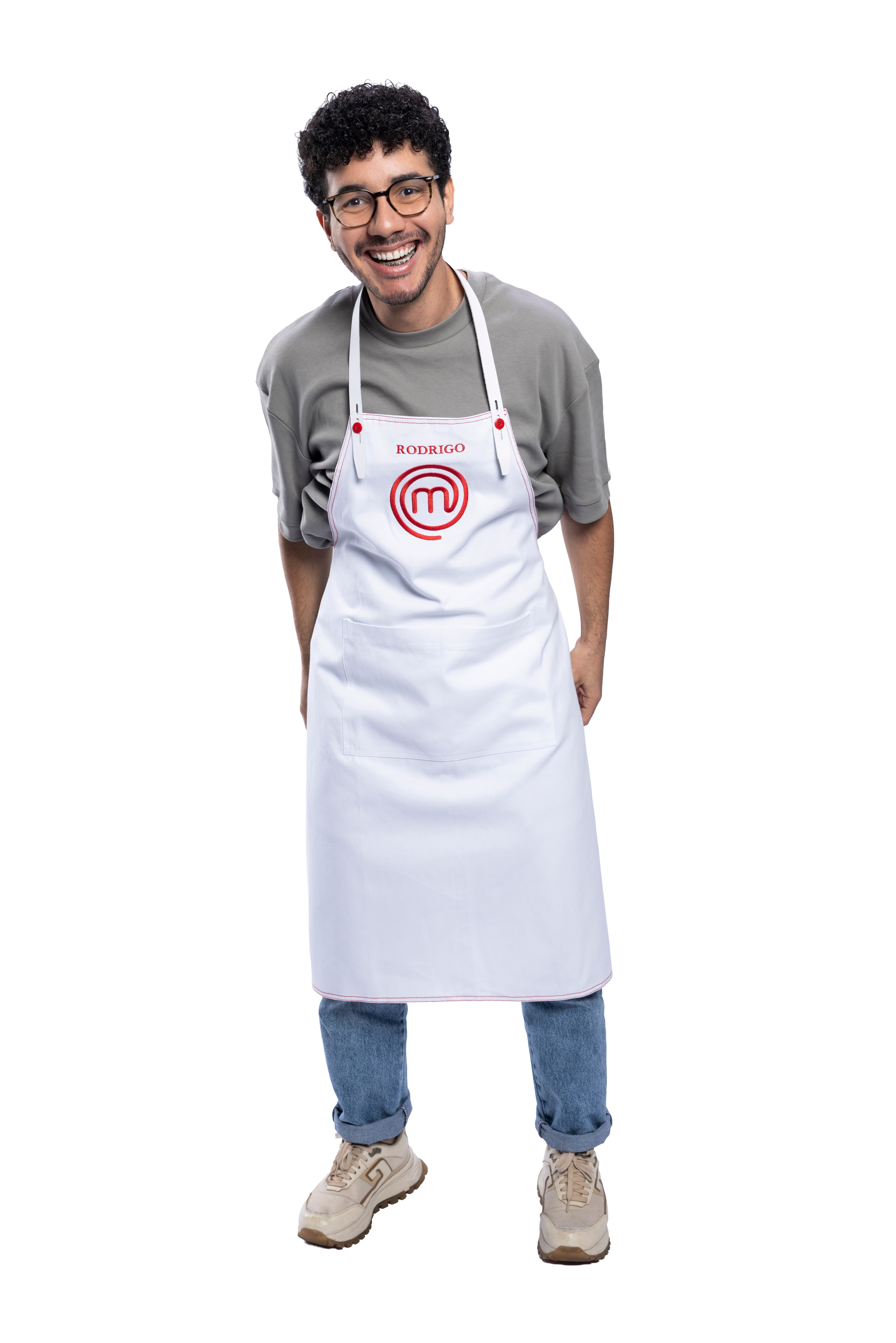 Homem jovem de pele clara, cabelo crespo escuro e &oacute;culos, sorrindo, vestindo camiseta cinza, jeans e t&ecirc;nis bege, com um avental branco do MasterChef escrito 