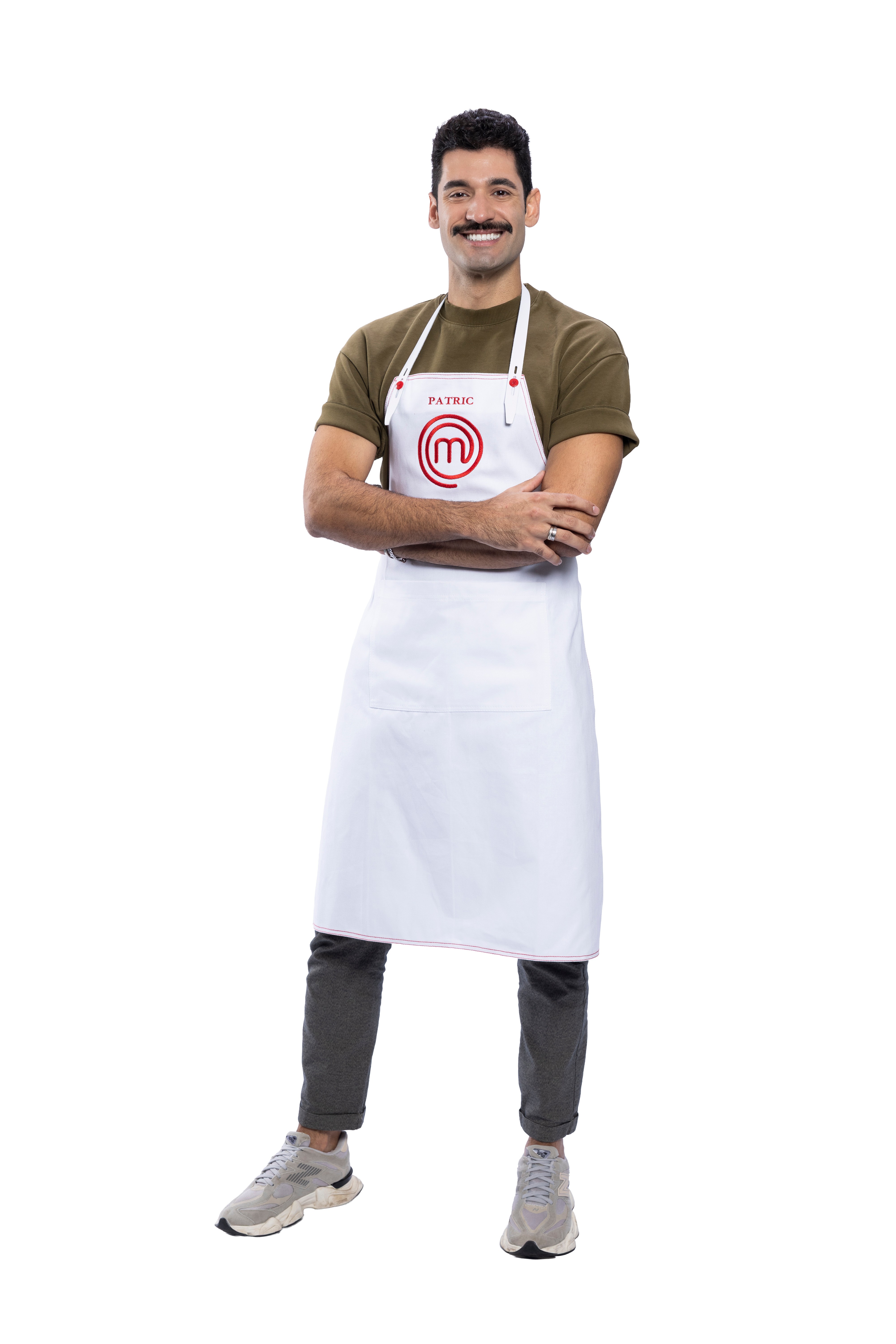 Homem sorridente com bigode, cabelo escuro e barba rala, vestindo camiseta verde-oliva, cal&ccedil;a cinza e t&ecirc;nis claros. Ele usa um avental branco com o nome 