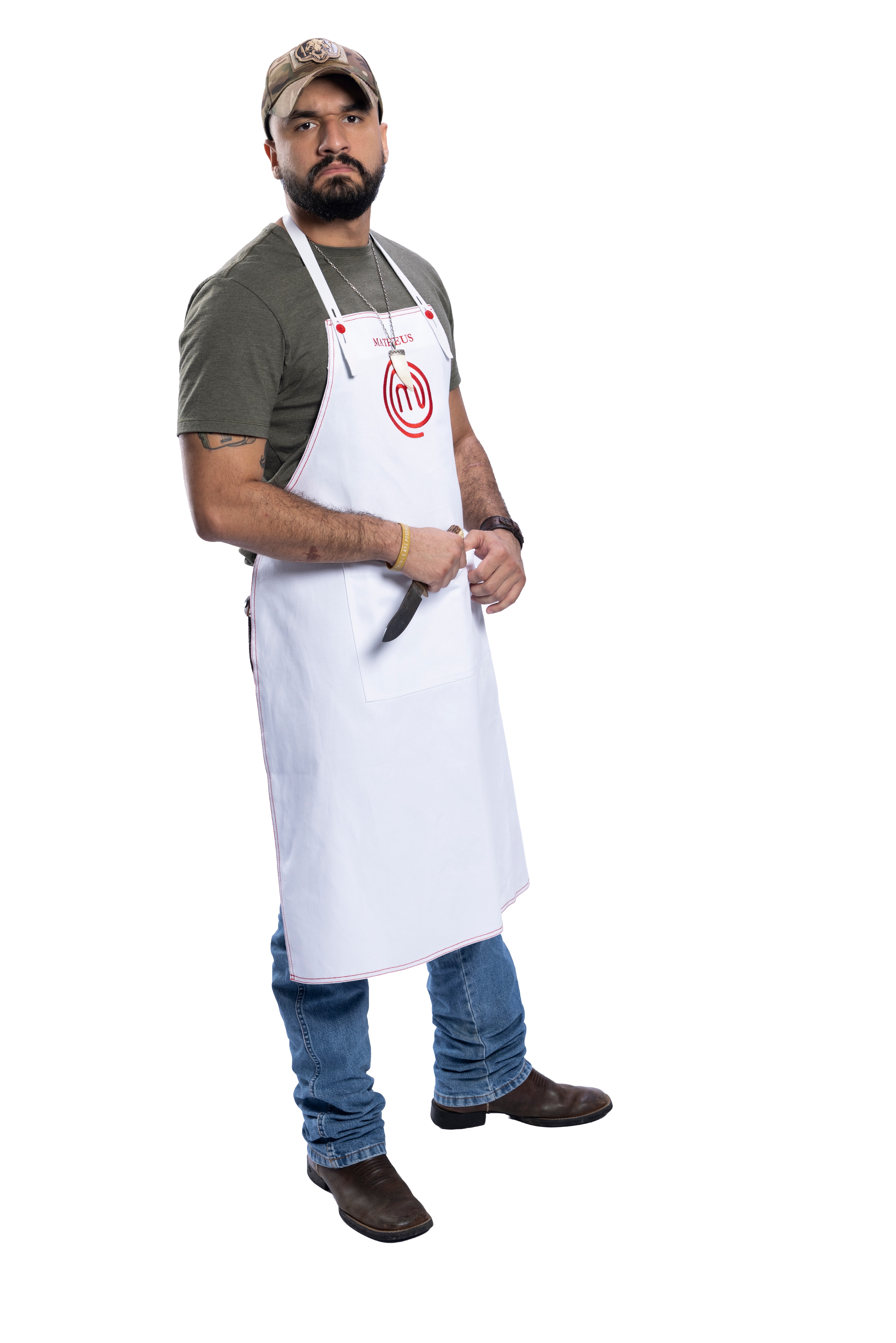 Homem barbudo, de bon&eacute; camuflado, camiseta verde-oliva e avental branco com logo MasterChef, segurando uma faca, jeans e botas marrons, em fundo branco