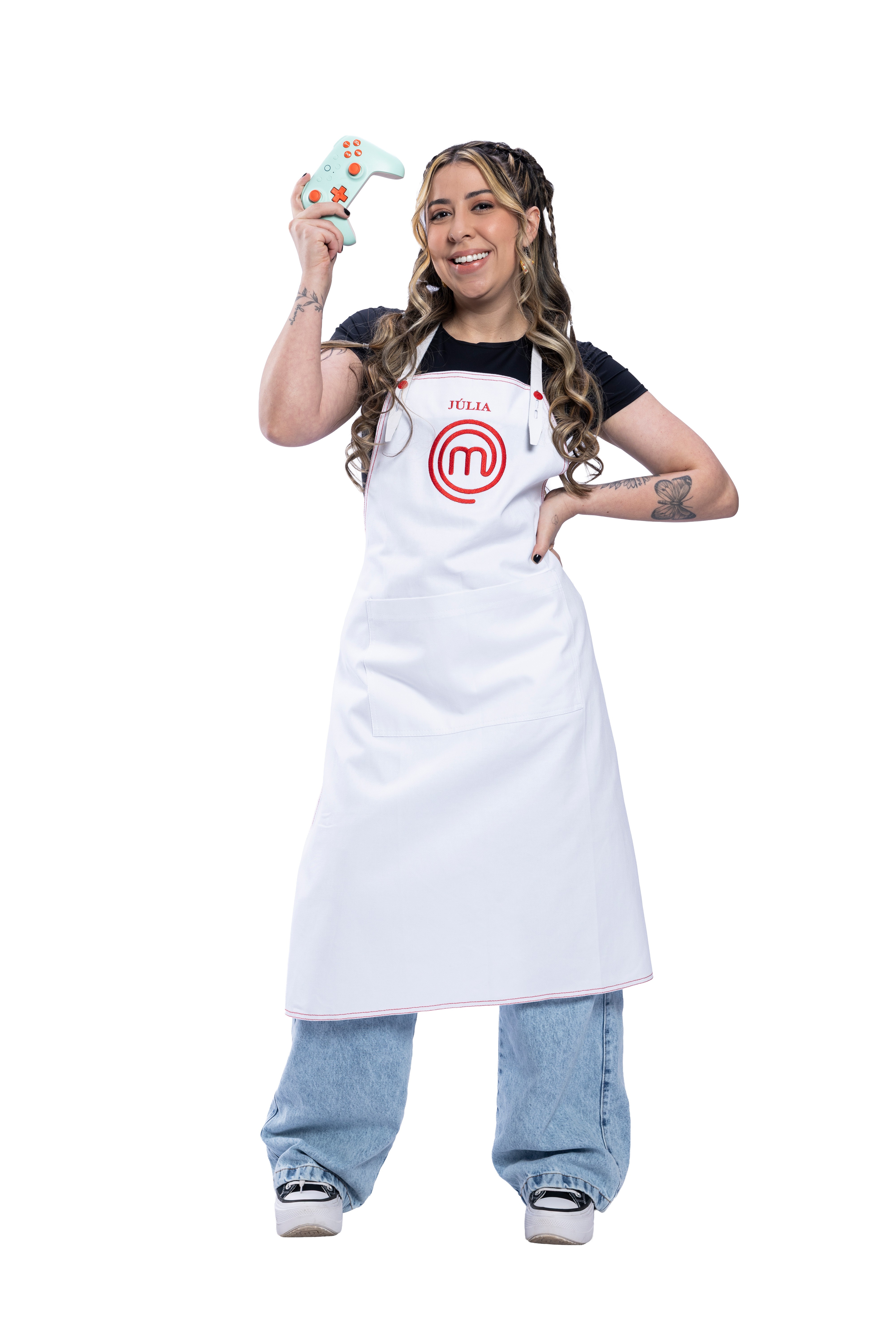J&uacute;lia, participante do MasterChef, sorri e segura um controle de videogame verde-&aacute;gua com detalhes laranja, vestindo avental branco com seu nome e o logo do programa, camiseta preta e cal&ccedil;a jeans larga